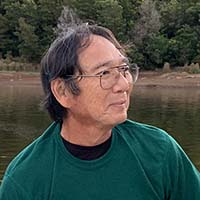 A person with glasses and dark hair, wearing a green shirt, is outdoors near a body of water with trees in the background.