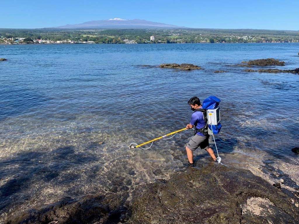 A person standing on rocky shoreline uses a long tool to sample shallow coastal water, wearing a backpack with equipment. Mountains, buildings, and trees are visible across the water under a clear blue sky.