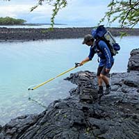 A person wearing a backpack stands on black volcanic rocks by the shore, using a long yellow pole to collect a sample from the calm, blue water under a cloudy sky.